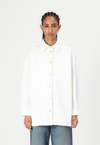 Cămașă albă oversized, cu nasturi, din bumbac, având guler clasic, cu nasturi din lemn și umeri căzuți, asortată cu blugi albaștri.