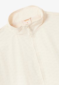 Veste zippée beige clair en tissu lisse ; dotée d'un col montant et d'un motif logo en relief subtil. L'intérieur a une doublure en mesh.