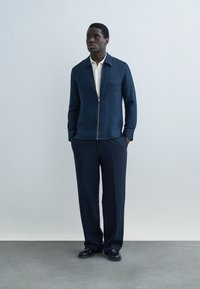 Moški stoji z rokami v žepih, oblečen v mornarsko modro jakno z zadrgo čez belo srajco, mornarsko hlače in črni čevlji na enostavni podlagi.