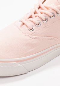 Světle růžové plátěné tenisky s kovovými očky, kulatými tkaničkami a bílou gumovou podrážkou mají jemný růžový pruh podél okraje podrážky.