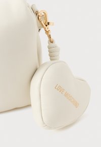Balta ādas rokassoma ar zelta aizdari. Pievienots ir balts sirds formas piekariņš ar tekstu "LOVE MOSCHINO" zelta krāsā.