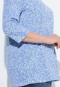 Blauw patroon top met driekwart mouwen, met een verspreide luipaardprint in donkerder blauw op een lichtblauwe achtergrond.