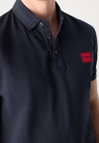 Granatowa koszulka polo z materiału bawełnianego. Posiada ściągaczowy kołnierzyk, zapięcie na trzy guziki oraz prostokątną naszywkę z logo "HUGO" w kolorze czerwonym.