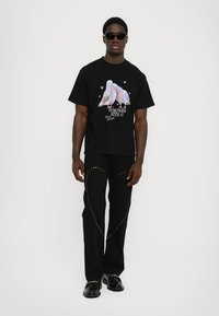 Svart t-shirt med ett grafik av två ljusa duvor och text. Svarta byxor med gyllene nitar i ett spiralformat mönster. Svarta skor.