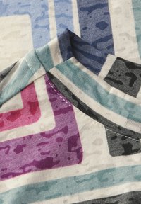 Monivärinen kangas, jossa on geometrisia kuvioita sinisen, vaaleanpunaisen, violetin, mustan ja valkoisen yhdistelmässä. Pehmeä rakenne, jossa näkyy saumoja ja ompeluyksityiskohtia.