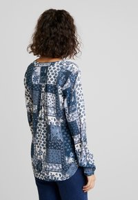 Chemise à manches longues avec un design patchwork en bleu et blanc, ornée de motifs floraux et cachemire. Tissu doux, encolure arrondie, et coupe décontractée.