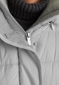 Grau gesteppte Jacke mit hohem Kragen, ausgestattet mit einem silbernen Reißverschluss, weichem Fleece-Futter und einem gerippten, dunklen Strickpullover darunter.