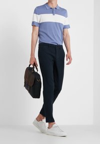 Muž oblečený v modro-bílem pruhovaném polo tričku, tmavých kalhotách, bílých teniskách, držící černou a hnědou aktovku, stojící na bílém pozadí.