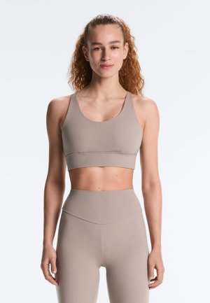 Kvinde med krøllet hår iført beige sports-bh og højtaljede leggings, stående oprejst mod en ensfarvet baggrund.