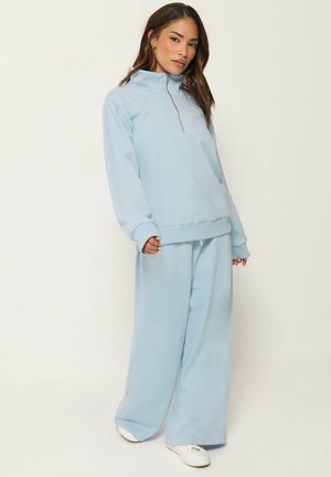 Donna che indossa una tuta azzurro chiaro, ampia, con top a collo alto con zip e pantaloni a gamba larga, in piedi davanti a uno sfondo semplice.