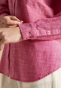 Ružová károvaná košeľa na gombíky s golierom a detailmi na manžetách. Obsahuje malé gombíky a mäkkú, textúrovanú látku. Spodný lem je zakrivený.