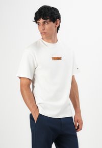 Muškarac koji nosi bijelu majicu s kratkim rukavima s narančastim logotipom "Tommy Jeans", plave hlače, stoji s rukama u džepovima, gleda lijevo.