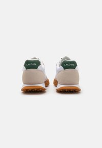 Witte sneakers met een groene hielaccent en beige suede, met een gestructureerde rubberen zool met spikes voor grip. Logo zichtbaar op de hiel.