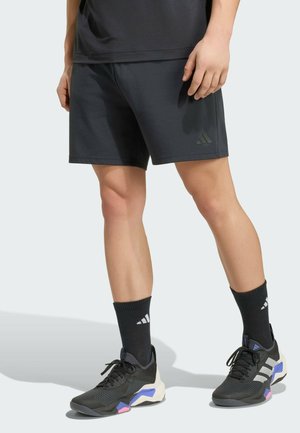 Osoba nosząca czarne krótkie spodenki sportowe, czarne skarpety Adidas oraz czarne buty sportowe z białymi, niebieskimi i różowymi akcentami, stojąca na jasnym tle.
