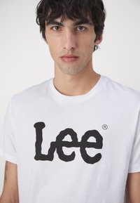Hvit bomull T-skjorte med rund hals og et stort, uregelmessig svart "Lee"-logo sentrert på brystet. Minimalistisk design, kortermet.
