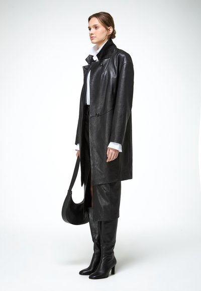 Frau in schwarzem Ledermantel, passendem Rock, kniehohen Stiefeln und weißem Hemd, hält eine schwarze Handtasche vor weißem Hintergrund.