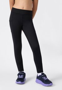 Femme portant des leggings noirs ajustés et des baskets noires et violettes, se tenant devant un fond blanc.