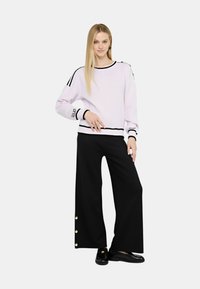 Maglione viola chiaro con accenti neri e maniche a strisce, abbinato a pantaloni neri a gamba larga con dettagli in bottone dorato. Scarpe nere.
