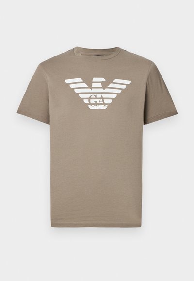 Kreminā t-kreklā ar apaļu kakla izgriezumu un īsām piedurknēm, uz kura ir balts ērgļa logo un svītru dizains krūšu daļā.