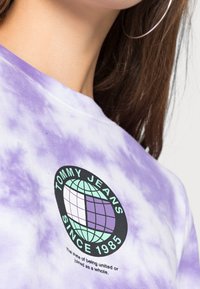 Fialové tričko s potiskem ve stylu tie-dye má kulatou černou nášivku se zeměkoulí a textem "TOMMY JEANS SINCE 1985" v bílých a mentolových akcentů. Bavlněný materiál.