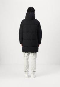 Abrigo acolchado negro con capucha, con un diseño de acolchado y un ajuste de longitud media. Lleva puesto sobre pantalones y zapatos de color claro.