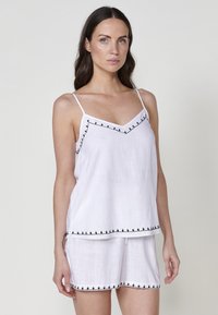 Witte linnen mouwloze top met verstelbare bandjes, voorzien van een v-hals en zwarte trim langs de randen. In combinatie met bijpassende shorts.