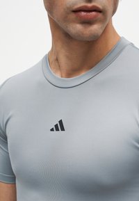 Gros plan sur une personne portant un t-shirt de sport gris clair avec un petit logo noir sur la poitrine.