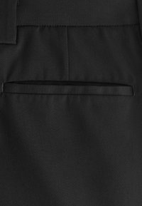 Tissu de pantalon noir sur mesure avec une poche passepoilée horizontale et un détail de ceinture montrant une texture de tissage diagonal subtile.