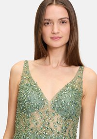 Robe de soirée verte avec décolleté en V et tissu translucide, ornée de broderies complexes en perles vertes et argentées, créant une texture scintillante.