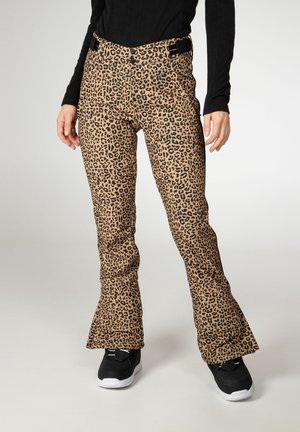 Persona que lleva pantalones acampanados con estampado de leopardo marrón y negro, zapatillas negras y una camiseta negra ajustada de manga larga, de pie contra un fondo liso.