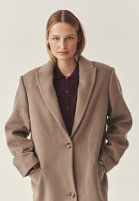 Beiger Wollmantel mit Reverskragen, Ein-Knopf-Verschluss und übergroßer Silhouette. Darüber getragen ein tiefbordeauxfarbenes Hemd mit Knöpfen.