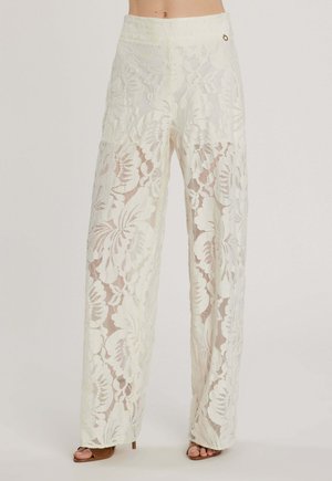 Pantaloni crema a vita alta con gamba larga e ampio motivo in pizzo floreale, indossati con tacchi aperti marroni, che mostrano l'addome e la parte inferiore del corpo.