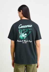 Camiseta de algodón negra con un gráfico de un esqueleto columpiándose, con "Converse" y "Parks & Wreck Dept. EST. 1908" impresos en blanco y verde.