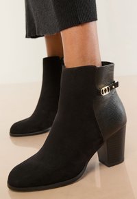 Ghete negre până la gleznă din piele intoarsă, cu un accent din piele ecologică, prevăzute cu o cataramă aurie și un toc masiv. Textură netedă, vârf ascuțit.