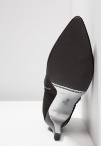 Black suede high-heeled shoe with a pointed toe, slim heel, and silver insole; features a small decorative zipper detail.