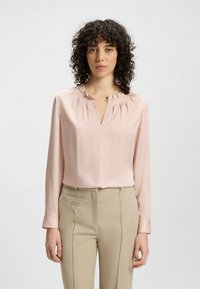Blusa a maniche lunghe di un delicato rosa pallido con un colletto arricciato e un dettaglio a oblò. Tessuto liscio, abbinata a pantaloni beige su misura con cuciture laterali.