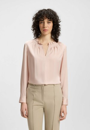 Blusa a maniche lunghe di un delicato rosa pallido con un colletto arricciato e un dettaglio a oblò. Tessuto liscio, abbinata a pantaloni beige su misura con cuciture laterali.