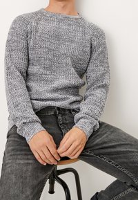 Grauer gestrickter Pullover mit strukturiertem Muster, rundem Ausschnitt und langen Ärmeln. Kombiniert mit schwarzen Jeans und auf einem Hocker sitzend.