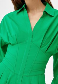 Kobieta w jasnozielonej sukience z głębokim dekoltem w kształcie litery V, z wyraźnie zarysowaną talią i długimi rękawami.