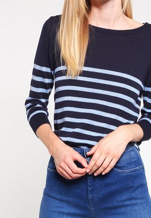 Persoon met lang blond haar draagt een marineblauwe trui met lichtblauwe horizontale strepen en maakt een knoop vast op een blauwe spijkerbroek.