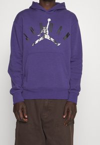 Sudadera con capucha morada que cuenta con un bolsillo canguro frontal y un gráfico en negro y plata del logo "Jumpman". Fabricada en una suave mezcla de algodón.