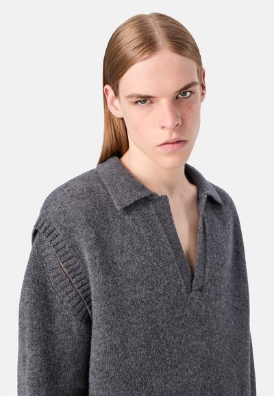 Junge Person mit langen, glatten blonden Haaren, die einen grauen Strickpullover mit tiefem V-Ausschnitt und ausschnitten an den Schultern trägt.