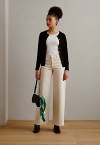 Zwarte button-up cardigan, witte geribde tanktop, beige wijde broek, zwarte handtas met een groene sjaal, zwarte enkellaarsjes, minimalistische stijl.