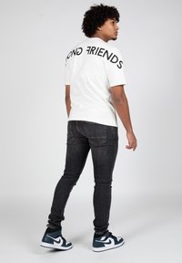 Camiseta blanca con letras negras que dice "AMIGOS FUERTES" en la parte de atrás, combinada con jeans negros y zapatillas Nike azules.
