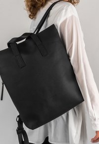 Czarny skórzany plecak o fakturowanej powierzchni, z dwoma górnymi uchwytami i odpinanym paskiem. Minimalistyczny design, bez widocznych logo i wzorów.