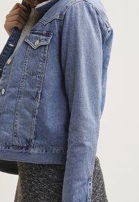 Giacca di jeans blu chiaro con design cropped, dotata di bottoni metallici, due tasche frontali e un colletto testurizzato.