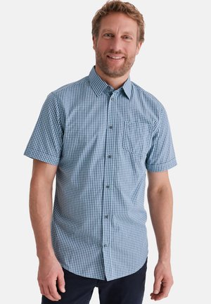 Lächelnder Mann mit kurzen lockigen Haaren, der ein blau kariertes Kurzarmhemd mit Knopfleiste und dunkle Hosen trägt, vor weißem Hintergrund stehend.