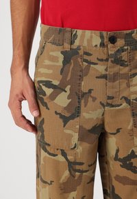 Camouflage cargo broek in tan en groene tinten, met een knoopsluiting, zijzakken en een gestructureerde stof. Hand rustend op de broek.