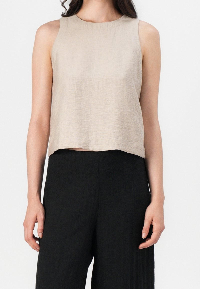 Beiges ärmelloses Oberteil mit strukturierter Oberfläche, rundem Halsausschnitt und leicht kurzem Schnitt, kombiniert mit schwarzen, weit geschnittenen Hosen.