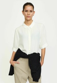 Bijela bluzica kratkih rukava s dugmadi sprijeda i suptilnom vezom na manžetama, uparena s bež hlačama i crnim puloverom vezanim oko struka.